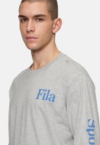T-shirt grigia a maniche lunghe con logo "Fila" blu sul petto e la scritta "sports" stampata sulla manica. Tessuto morbido e vestibilità comoda.