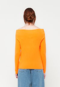 Pulover z oranžno pletenino, s spušenimi rameni in dolgimi rokavi, z rebrasto strukturo in oprijetim krojem, v kombinaciji s svetlo modrimi širokimi hlačami.