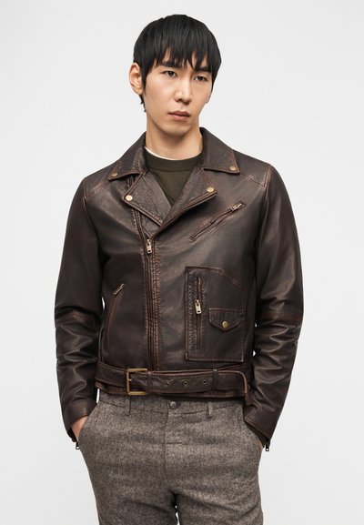Νέος άνδρας που φοράει σκούρο καφέ δερμάτινο μπουφάν μοτοσικλετιστή με φερμουάρ και λεπτομέρειες με πόρπες, συνδυασμένο με γκρι παντελόνια υφής και πράσινο πουλόβερ.
