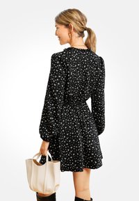 Donna con un ponytail biondo che indossa un vestito floreale nero a maniche lunghe, che tiene in mano una borsa beige, vista di spalle e sorridente.