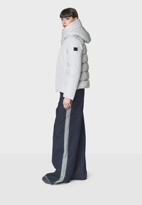 Giacca piumino grigia chiara con colletto alto e tasche laterali, abbinata a jeans a gamba larga blu scuro con dettaglio a striscia laterale.