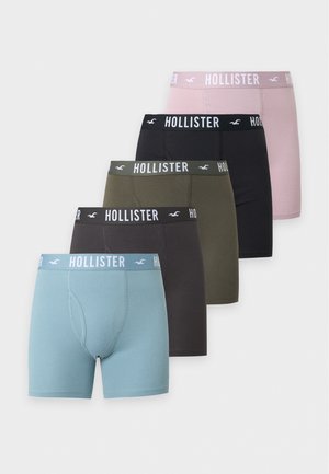 Homme debout, pieds nus, portant un boxer Hollister gris foncé, regardant sur le côté sur un fond blanc uni.