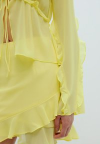 Geel chiffon jurk met gelaagde ruffles langs de mouwen en de zoom. Strikjes bij de halslijn zorgen voor extra detail in de lichte stof.