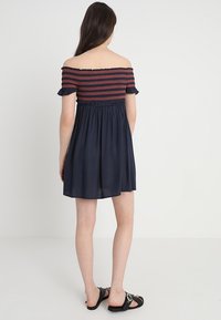 Ung kvinna som står vänd bort med en off-shoulder marinblå klänning och rödrandig topp samt svarta sandaler på en vanlig vit bakgrund.