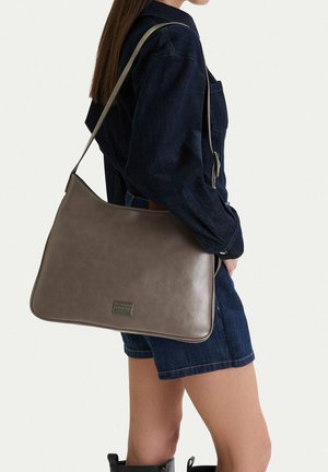 Mujer con vestido oscuro de mezclilla y botas negras, llevando un gran bolso de cuero taupe al hombro con una etiqueta de marca visible.