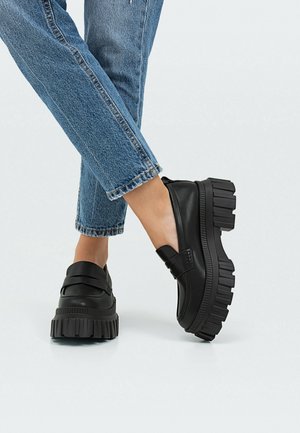 Zwarte leren plateauzolen met een dikke zool, voorzien van een gekruist bandontwerp en een gladde textuur, gecombineerd met blauwe denim.