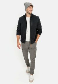 Donkergrijze rits-fleecejack, witte t-shirt, tan cargobroek, grijze beanie en lichte sneakers; voorzien van een ritszak en cargozakken.