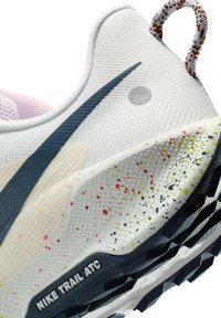 Gros plan sur le talon d'une chaussure Nike Trail ATC blanche avec un logo Swoosh noir, une semelle intermédiaire multicolore tachetée, une semelle texturée et une languette de tirage rouge et blanche.