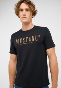 Czarna koszulka z krótkim rękawem z miękkiej tkaniny, z wyraźnym złotym logo "MUSTANG" oraz napisem "JEANS COMPANY SINCE 1932" w casualowej czcionce.