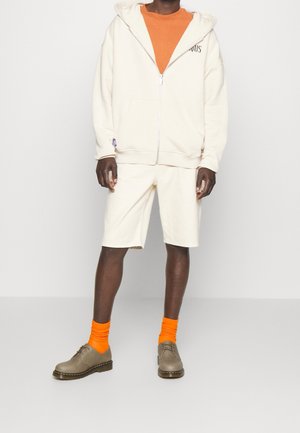 Felpa con zip color crema, t-shirt arancione, pantaloni corti color crema, calzini arancioni, scarpe grigie con suola nera. Stile casual, vestibilità rilassata, tessuto texturizzato.