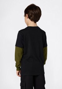Ein Kind mit kurzen braunen Haaren trägt ein schwarzes kurzärmliges Shirt über einem olivgrünen Langarmshirt und schwarzen Cargo-Hosen, von hinten gezeigt.
