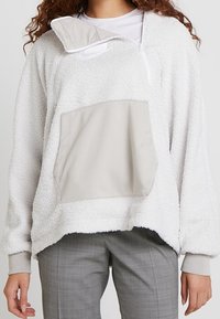 Bílý fleecový pulovr s polovičním zipem u límce, vybavený velkou hladkou světle šedou přední kapsou a žebrovanými manžetami.