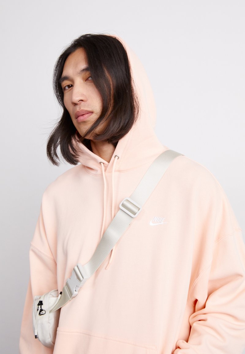 Różowa bluza z kapturem, sznurkami i przednią kieszenią, z białym logo Nike. W połączeniu z beżową torebką na ramię. Gładka tekstura materiału.