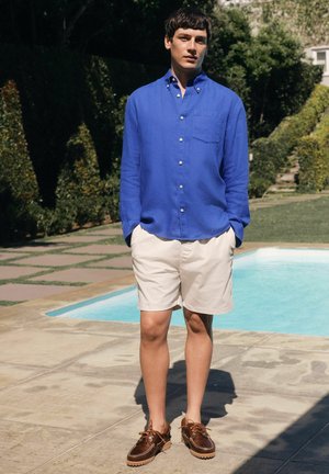 Mand står ved poolen iført en klar blå skjorte med knapper, beige shorts og brune lædersko med snørebånd i en solrig have.