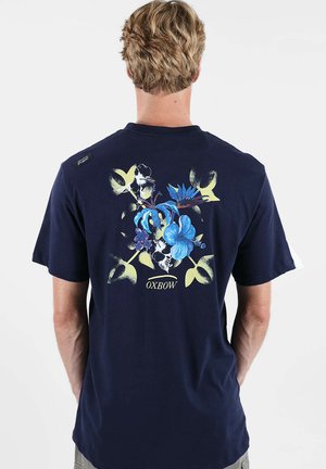 T-shirt bleu marine à manches courtes avec un motif floral bleu et jaune au dos, agrémenté d'un accent noir et d'une étiquette logo sur l'épaule.
