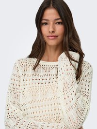 Giovane donna con lunghi capelli scuri indossa un maglione bianco lavorato a maglia con motivi traforati, posando con la mano che tocca i capelli su uno sfondo semplice.