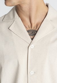 Camisa de botones color beige claro, hecha de tela texturizada, con cuello tipo camp, dos botones y un tatuaje negro visible debajo.