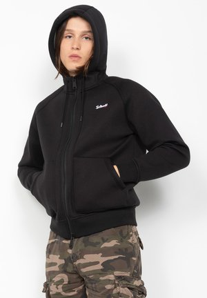 Ung person iført sort hættetrøje med lynlås og camouflage cargo-bukser, hænderne i jakkelommerne, vendt fremad mod en hvid baggrund.