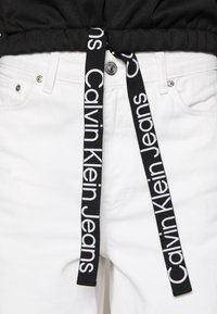 Černý látkový pásek s logem "Calvin Klein Jeans", připevněný k bílým džínovým šortkám s kovovým knoflíkem a kapsami, plochá textura.