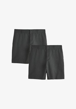 Homme debout devant un fond blanc, portant un polo gris clair à manches courtes, un short gris foncé, des chaussettes claires et des baskets blanches.