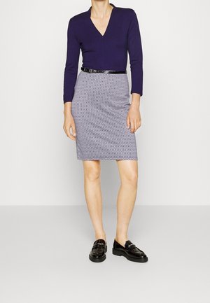 Vrouw draagt een marineblauw V-hals langermig shirt, een knielange rok met patroon, een zwarte riem en zwarte loafers, staande tegen een effen achtergrond.