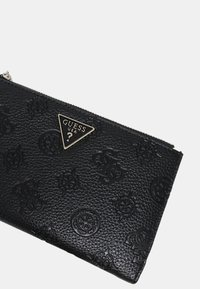 Pochette zippée noire texturée avec des motifs en relief et une plaque en métal triangulaire portant l'inscription "GUESS USA ?" près du bord supérieur.