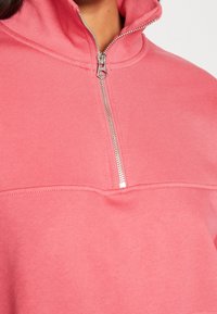 Světle růžová mikina na zip vyrobená z měkké tkaniny, s vysokým límcem a stříbrným zipem, s hladkou texturou a minimálním švem.
