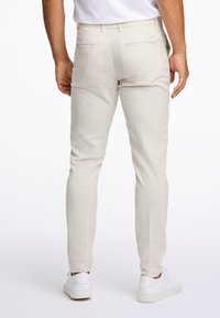 Mężczyzna w dopasowanych beżowych spodniach i białych trampkach, stojący z rękami swobodnie opuszczonymi wzdłuż ciała na białym tle.