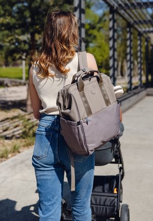 Gray and beige backpack with multiple compartments, adjustable straps, and a textured lower section, carried by a person in denim jeans.