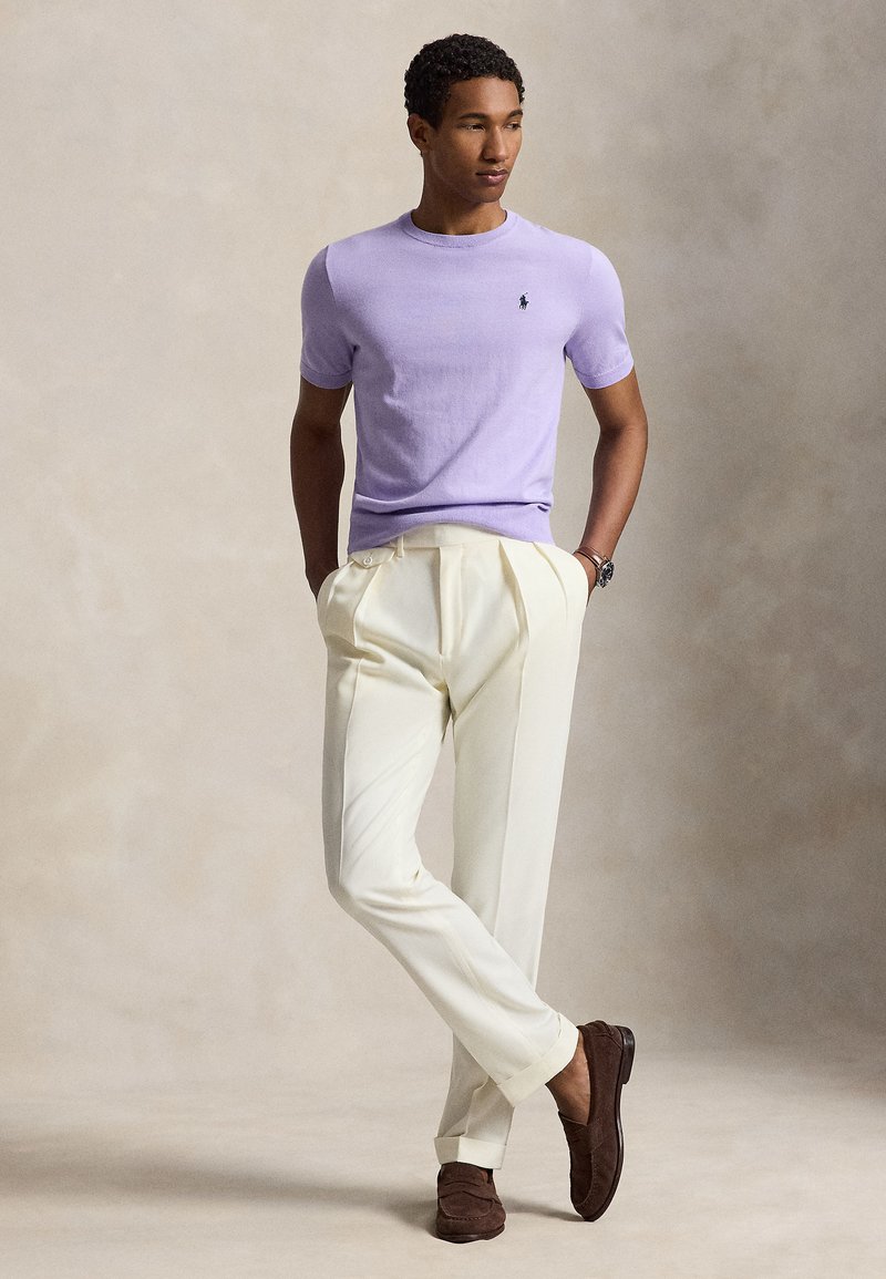 Un homme debout, les mains dans les poches, portant un haut en maille lavande à manches courtes, un pantalon plissé crème et des mocassins marron sur un fond neutre.