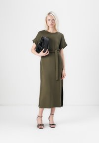 Olivgrön, midiklänning med bälte, korta ärmar och sidofickor, i kombination med en svart handväska och svarta klacksandaler med polka prickar.