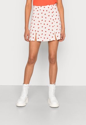 Kobieta ubrana w białą mini spódnicę z czerwonym nadrukiem truskawek i dwoma przednimi rozcięciami, pomarańczową bluzkę oraz białe botki na sznurowadła.