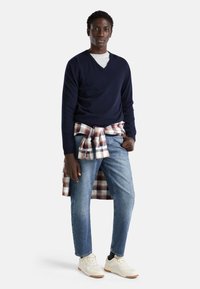 Granatowy sweter w serek noszony na szarym t-shircie, w połączeniu z jasnoniebieskimi dżinsami. W kratkę koszula przewiązana w talii. Białe sneakersy.