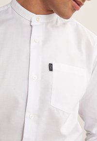 Hvid skjorte med ståkrave, knaplukning foran og en brystlomme på venstre side. Stoffet virker glat med en let glans; har et lille logomærke.