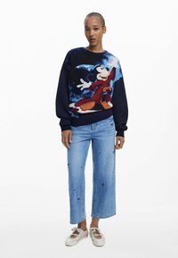 Strickpullover mit einem blau-schwarzen Design und einer Mickey-Maus-Grafik; kombiniert mit hellblauen Jeans mit verstreuten schwarzen Akzenten.