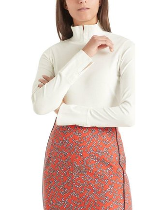 Femme portant un haut blanc à manches longues avec un col haut et une jupe orange avec un petit motif floral, posant les bras croisés.