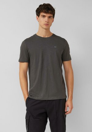 Młody mężczyzna w ciemnoszarej koszulce z krótkim rękawem i czarnych spodniach bojówkach, stoi na jasnym, jednolitym tle.