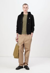 Fato com capuz preto fechado sobre uma camisa curta de manga oliva, calças soltas bege, sapatos pretos, segurando uma mala bege. Estilo minimalista, texturas simples.