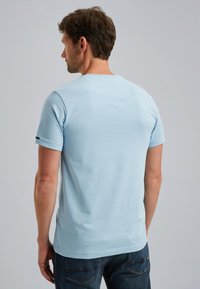 T-shirt en coton bleu clair à manches courtes, avec un patch rectangulaire au col arrière et un petit logo sur la manche gauche.