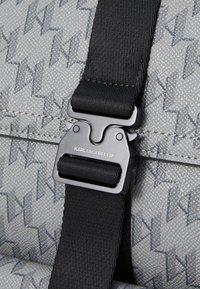 Sac gris au fini texturé, doté d'une sangle noire et d'une boucle en métal argenté ajustable gravée "KARL LAGERFELD."