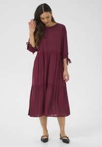 Burgundy midi-klänning med lagerdesign, trekvartsärmar och knyt detaljer vid manschetterna. Tillverkad av lätt, flödande tyg.