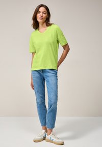 T-shirt vert vif à manches courtes et col en V. Associé à un jean bleu clair à rayures verticales et des baskets blanches avec des accents bleus.