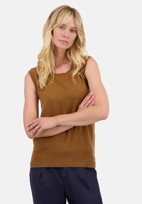 Haut tricoté marron sans manches avec un col rond, coupe droite et ourlet côtelé. Le modèle a les bras croisés, portant un pantalon bleu marine. Texture unie.