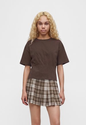 Jeune femme aux longs cheveux blonds bouclés porte un haut marron à manches courtes et une jupe plissée à carreaux marron et blanc, debout devant un fond blanc.