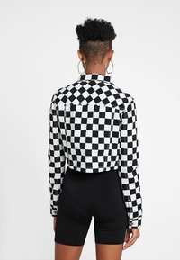 Person med kort, krulligt hår som bär stora hexagonala hoopörhängen, en svartvit rutmönstrad jacka och svarta cykelshorts, vänd bort.