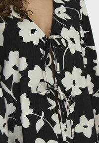 Schwarze Bluse mit floralem Wickeldesign, großen weißen Blumenmustern, einem tiefen V-Ausschnitt und einer Bindeschließe am Hals mit kontrastierenden Bindebändern.