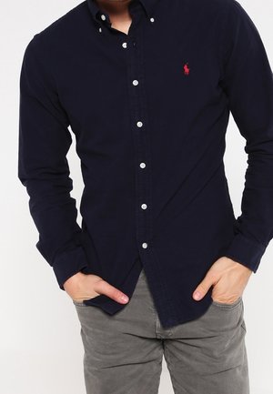 Man draagt een donkerblauw overhemd met knopen en grijze broek met handen gedeeltelijk in de zakken tegen een effen witte achtergrond.