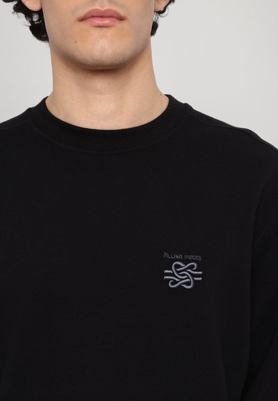 Sweatshirt noir avec un col rond, présentant un logo gris subtil et un design de nœud sur la poitrine, fabriqué dans un tissu doux avec une texture lisse.