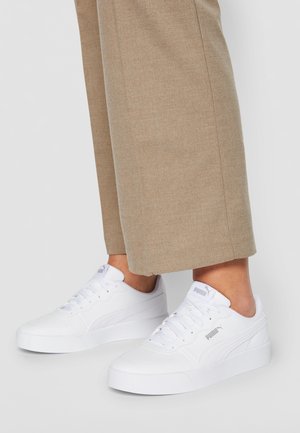 Weiße Puma-Sneaker mit Schnürsenkeln, getragen zu beigen Hosen, von Knöchel bis zur Sohle auf grauem Hintergrund dargestellt.