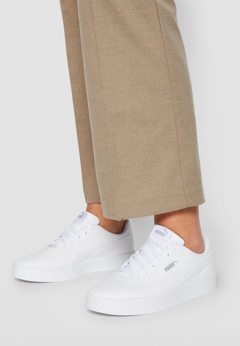 Baskets blanches Puma avec lacets portées avec un pantalon beige, montrées de la cheville à la semelle sur fond gris.